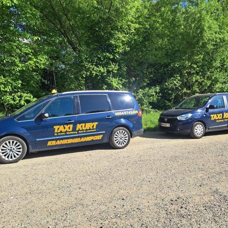 Zwei blaue Taxis auf einem Kiesweg mit Bäumen im Hintergrund geparkt. Ein Taxi hat die Aufschrift 'TAXI KURT' an der Seite.