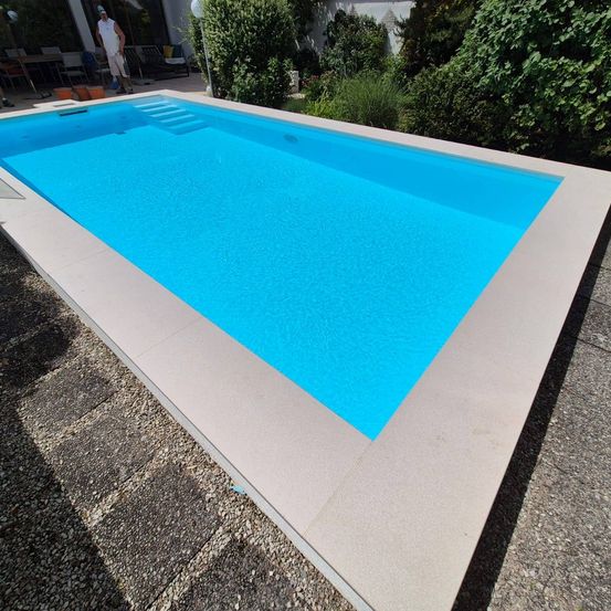 Ein leerer rechteckiger Swimmingpool mit blauer Wasseroberfläche und hellgrauem Pooldeck, umgeben von Kies und Grün.