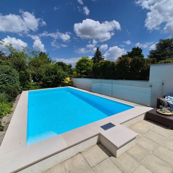 Ein klarer blauer Swimmingpool steht in einem Hinterhof, umgeben von Grün und einer weißen Umzäunung unter einem blauen Himmel.