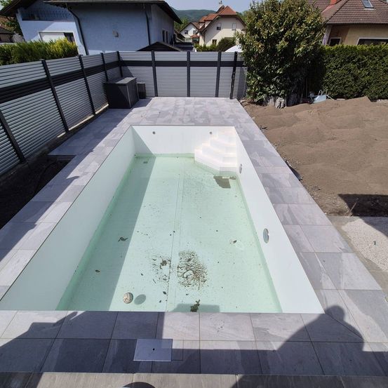 Ein leerer rechteckiger Pool mit gefliesten Kanten steht in einem Hinterhof, umgeben von einem Zaun und einem Haus im Hintergrund.