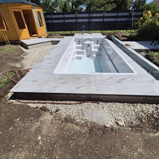 Ein Außenwhirlpool, teilweise mit Wasser gefüllt, befindet sich im Aufbau in einem Garten. Es gibt ein Holzhaus und eine Steinmauer.