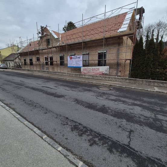Ein Ziegelgebäude im Bau mit Gerüsten an der Straßenseite. Ein Banner mit dem Text 'Meitz-Dach' ist am Gebäude befestigt.