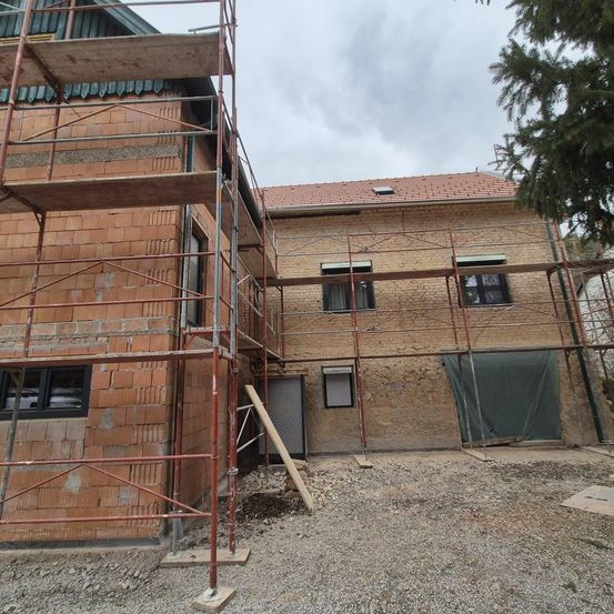 Ein Haus aus Ziegelsteinen im Bau mit Gerüsten drumherum. Die Außenwände sind aus Ziegelsteinen und es gibt einen großen Baum in der Nähe.