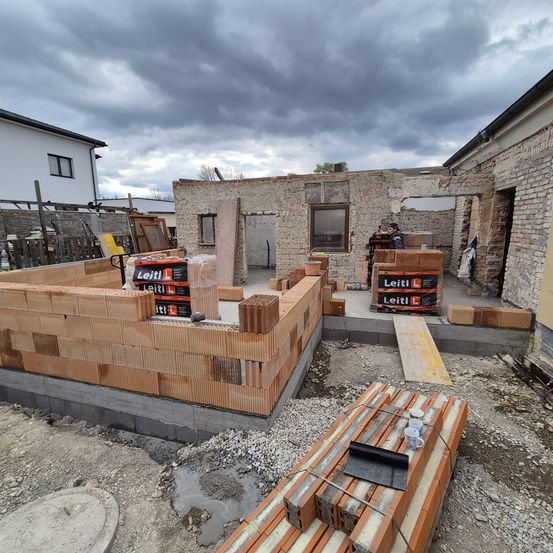 Ein Gebäude im Bau mit bewölktem Himmel. Stapel von Ziegeln, Lagerboxen und eine Rampe sind sichtbar. Eine Person steht in der Nähe des Gebäudes.