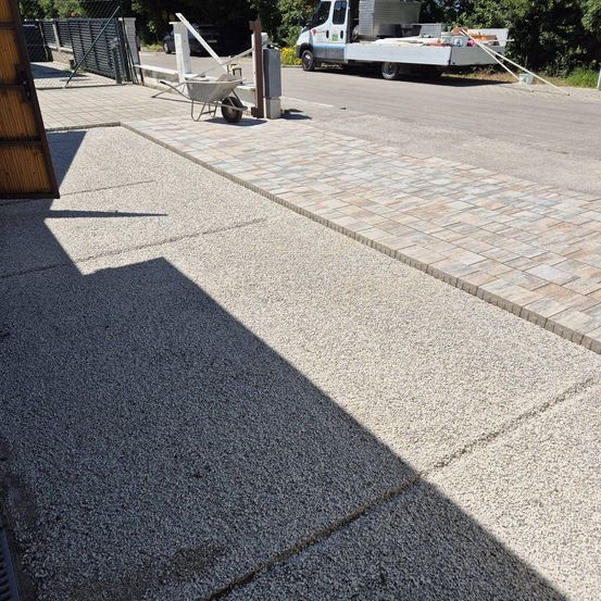 Eine Einfahrt mit einer Ziegelbordüre und einem geparkten Lieferwagen daneben. Ein Schubkarren steht auf dem Ziegelabschnitt. Im Hintergrund sind Bäume zu sehen.
