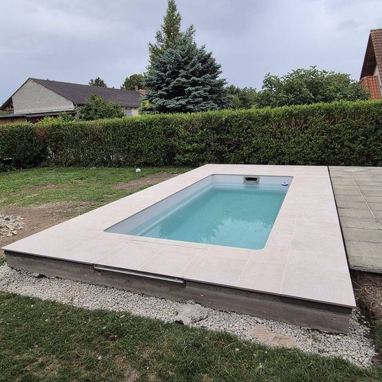 Ein leerer Pool, umgeben von einer Steinumrandung, befindet sich in der Mitte des Rasenbereichs. Hinter dem Pool steht eine grüne Hecke. In der Ferne ist ein Haus mit einem braunen Dach zu sehen.