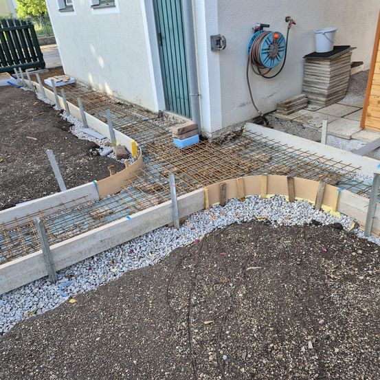 Bauarbeiten finden vor einem weißen Haus statt. Ein Betonweg mit Kies wird gebaut. Eine grüne Gartenschlauch ist an einer Wand angeschlossen.