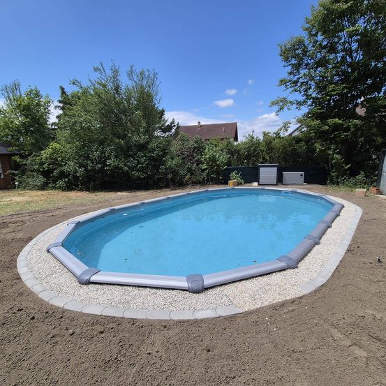 Ein ovaler Pool ist im Garten installiert, umgeben von einem weißen Kiesrand. Der Pool ist mit klarem Wasser gefüllt. Bäume und ein Haus sind im Hintergrund.