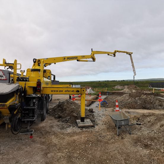 Ein gelber Baufahrzeug mit 'Pehofer' darauf steht über einer Baustelle, mit einem Schubkarren in der Nähe. Orangefarbene Verkehrskegel sind um das Gebiet platziert.
