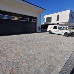 Ein weißer Van ist vor einem modernen Haus mit einer gepflasterten Einfahrt, einer schwarzen Garagentür und Glasfenstern geparkt. Der Himmel ist klar.