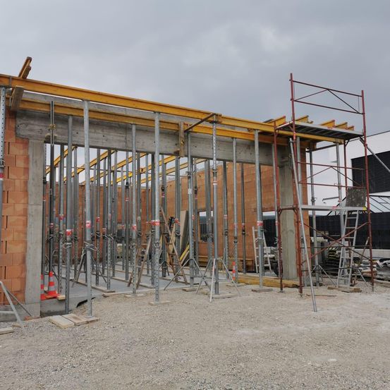 Eine Baustelle unter einem bewölkten Himmel mit einer teilweise fertiggestellten Ziegelmauer, Metallgerüst und einem gelben Stahlträger.