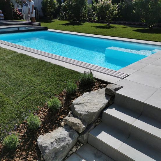 Ein sauberer und klarer Swimmingpool, umgeben von einem üppigen grünen Rasen mit Steinen und Pflanzen, unter einem sonnigen Himmel.