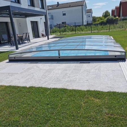 Ein überdachter Pool befindet sich auf einer Betonterrasse mit einem modernen Haus im Hintergrund. Die Terrasse hat einen Esstisch mit Stühlen. Der Pool ist mit einem transparenten Dach bedeckt.