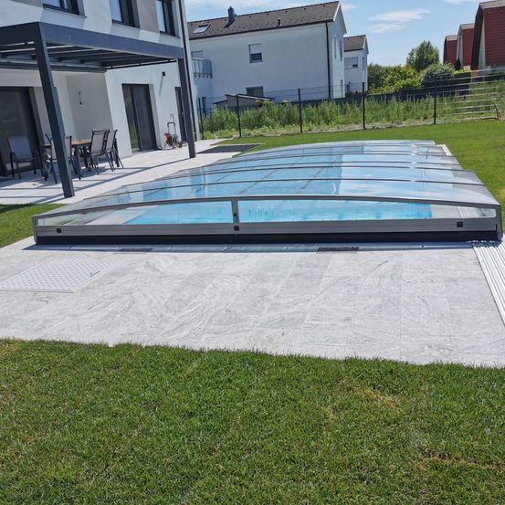 Ein überdachter Pool befindet sich auf einer Betonterrasse mit einem modernen Haus im Hintergrund. Die Terrasse hat einen Esstisch mit Stühlen. Der Pool ist mit einem transparenten Dach bedeckt.