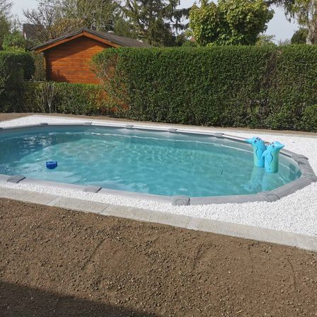 Ein rechteckiger Pool steht im Freien, mit einem grauen Rand und klarem blauem Wasser. Zwei blaue Spielzeuge schwimmen im Wasser. Hinter dem Pool steht ein Holzhaus und grüne Hecken.