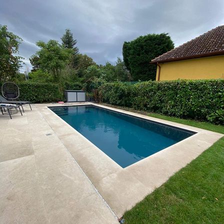 Ein moderner rechteckiger Pool ist von einer gefliesten Terrasse umgeben, mit einem gelben Haus und Hecken. Zwei Liegestühle stehen auf der Terrasse. Bäume und Büsche im Hintergrund.