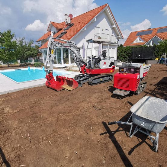 Ein roter Bagger steht neben einem Swimmingpool und einer weißen Schubkarre vor einem Haus mit rotem Dach.