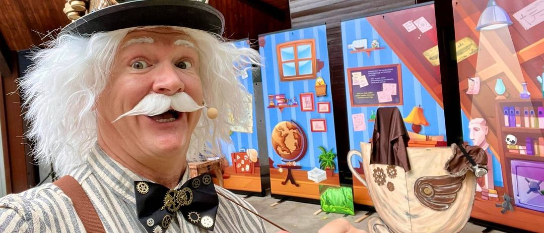 Ein Mann mit weißem Bart und Schnurrbart, der einen Steampunk-Hut und eine Fliege trägt, steht vor farbenfrohen Plakaten.
