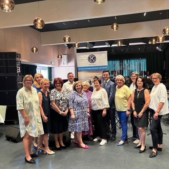 Eine Gruppe von Menschen posiert für ein Foto in einem Saal, einige tragen Brillen und Halsketten, mit einem Banner und Lautsprechern im Hintergrund.