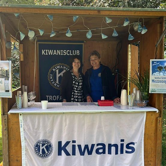 Zwei Frauen stehen vor einem hölzernen Kiosk mit einem Schild, das 'Kiwanis Club' besagt. Der Kiosk ist mit Lichtern dekoriert und hat einen Tisch mit Tassen und einer Pflanze.