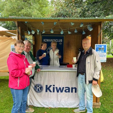 Fünf Personen stehen an einem Kiwani-Stand in einer Außenumgebung. Sie halten Tassen und lächeln. Der Stand hat ein weißes Tuch mit dem Wort Kiwan und einem Banner.