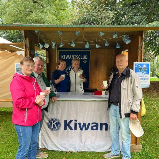 Fünf Personen stehen an einem Kiwani-Stand in einer Außenumgebung. Sie halten Tassen und lächeln. Der Stand hat ein weißes Tuch mit dem Wort Kiwan und einem Banner.