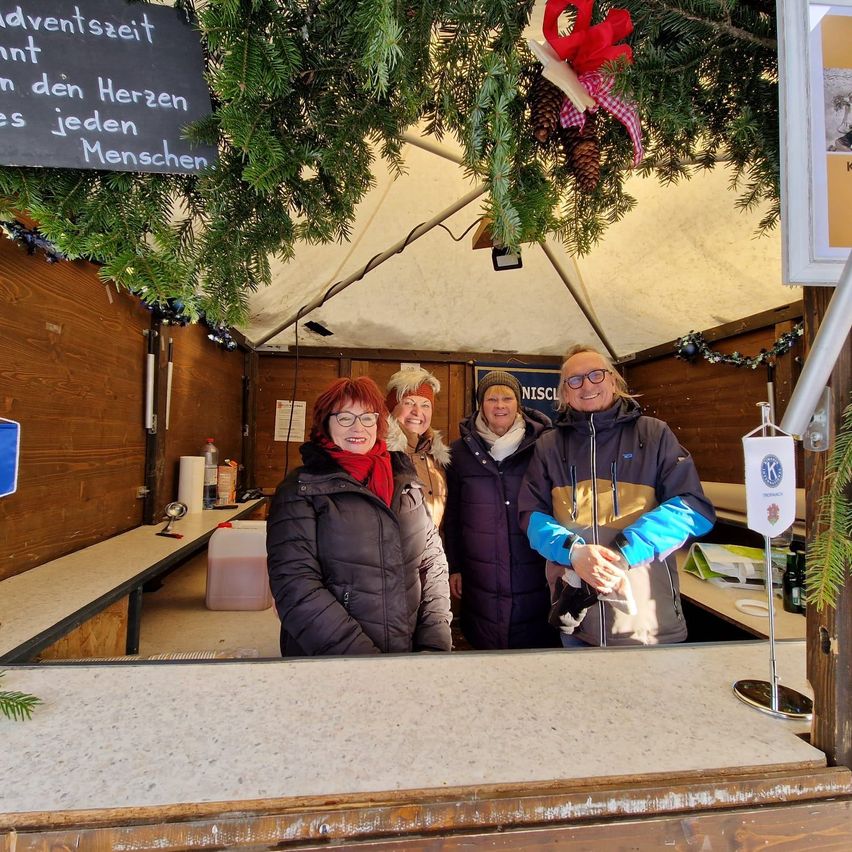 Vier Personen, warm gekleidet, stehen hinter einem Tresen in einer festlich geschmückten, holzverkleideten Umgebung. Oben steht ein Schild mit 'Adventszeit in den Herzen jedes Menschen', mit Tannenzapfen und Schleifen. Ein festlicher Baum hängt über ihnen.