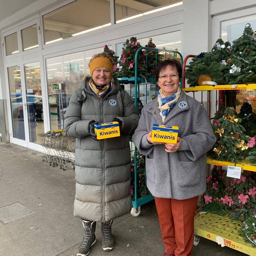 Zwei Frauen in Wintermänteln stehen vor einem Geschäft und halten Boxen mit der Aufschrift Kiwianis. Im Hintergrund befinden sich Weihnachtsbäume und Dekorationen.