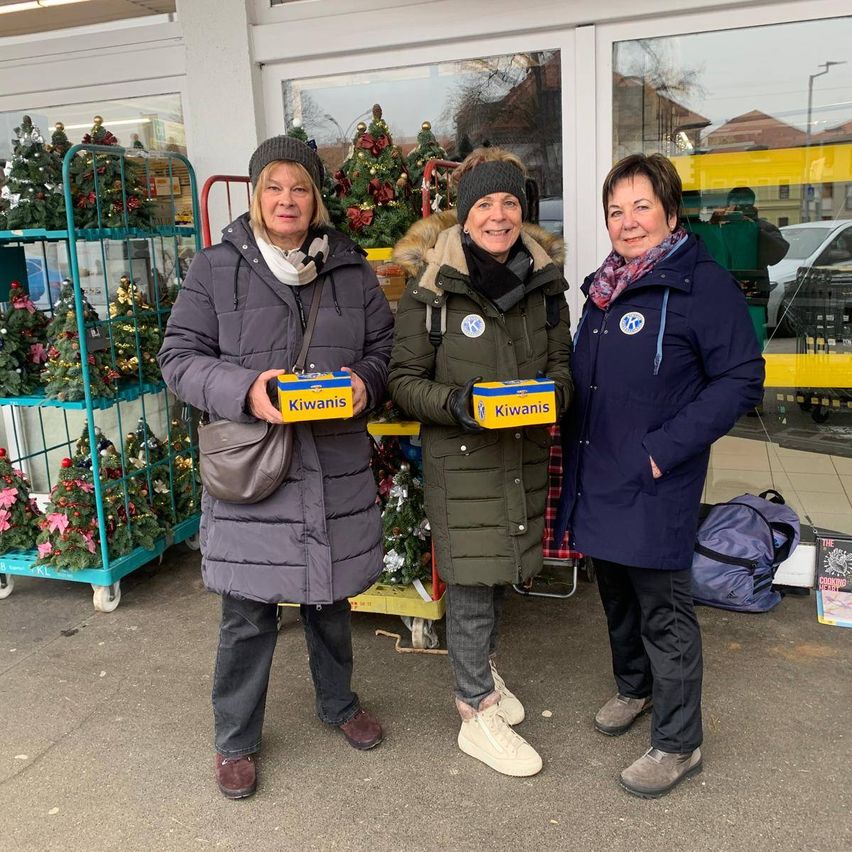 Drei Frauen stehen lächelnd für ein Foto draußen. Sie halten Schachteln mit der Aufschrift 'Kiwanis'. Eine trägt einen Schal, eine andere eine Mütze. Hinter ihnen steht ein Weihnachtsbaum und ein Trolley.