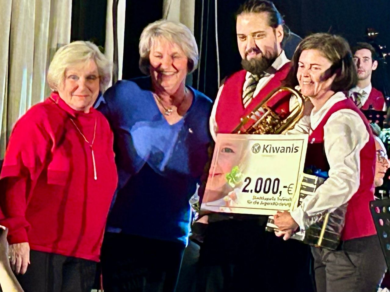 Vier Personen stehen eng beieinander und lächeln für ein Foto. Die Frau links trägt ein rotes Shirt, während die Frau neben ihr ein blaues Shirt trägt. Ein Mann im roten Weste hält eine Trompete. Ein gerahmtes Bild mit einem Baby wird mit einem Spendenbetrag angezeigt.