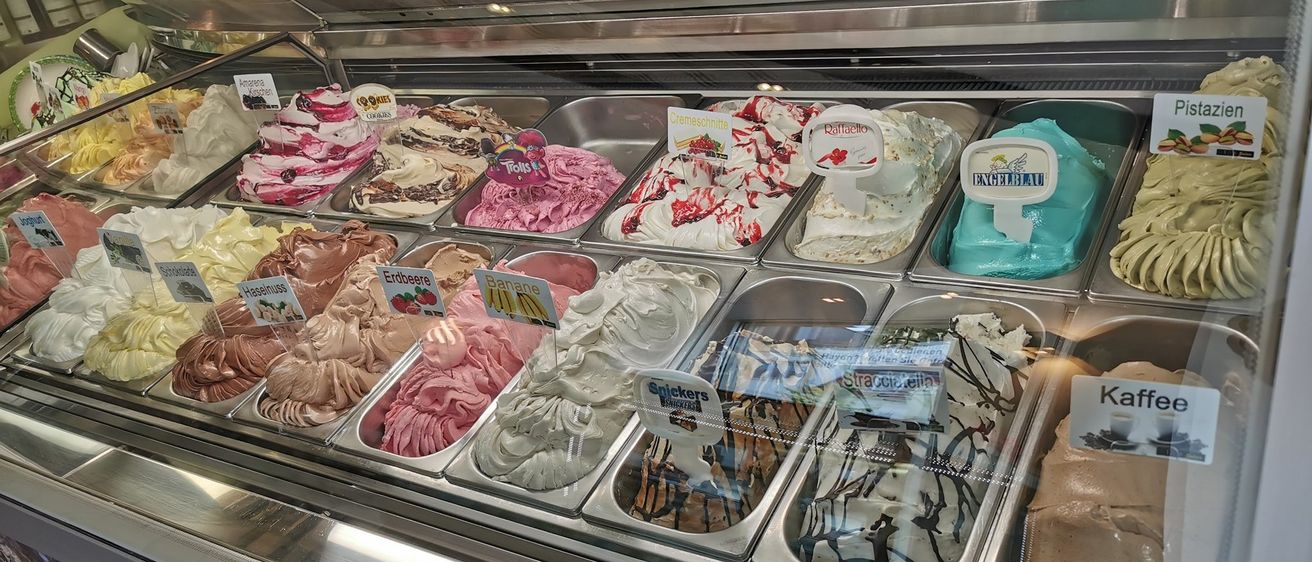 Eine Vitrine gefüllt mit verschiedenen Eissorten in unterschiedlichen Farben und Toppings. Jede Sorte hat ein Etikett, darunter 'Banana,' 'Stracci' und 'Kaffee.' Die Vitrine ist gut beleuchtet und zeigt die cremigen Texturen und Dekorationen.