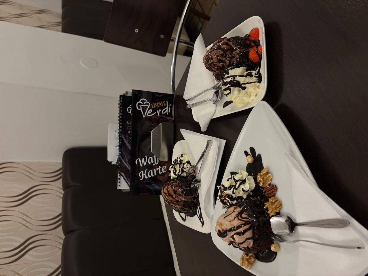 Drei Dessertteller mit Eiscreme und Toppings sind auf einem dunklen Tisch angeordnet. Jeder Teller hat eine andere Art von Eiscreme, garniert mit Schokoladensauce, Schlagsahne und Nüssen. Ein Menübuch und ein silberner Eiscremespachtel liegen in der Nähe.