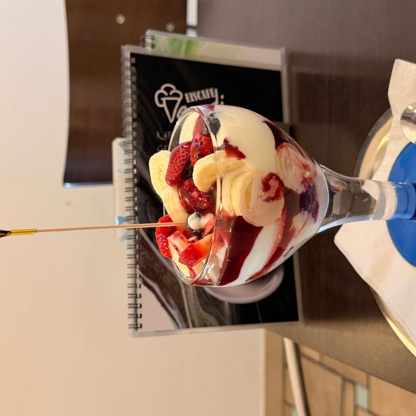 Ein Dessert in einem Weinglas gefüllt mit Joghurt, Erdbeeren, Bananen und einer Kirsche obenauf.