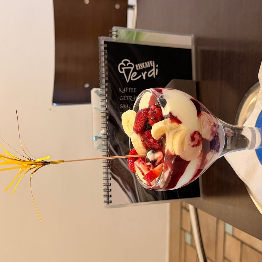 Ein Dessert in einem hohen Glas, mit Schichten aus Sahne, Beeren und Bananen, gekrönt von einem Strohhalm und einer festlichen Dekoration.