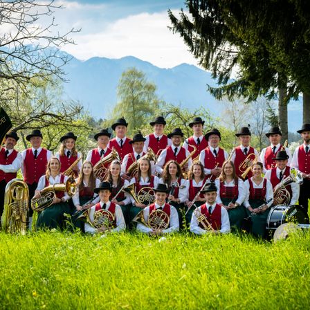 Eine große Gruppe von Musikern in roten und weißen Westen und schwarzen Hüten posiert auf einer Grasfläche mit Bergen im Hintergrund.