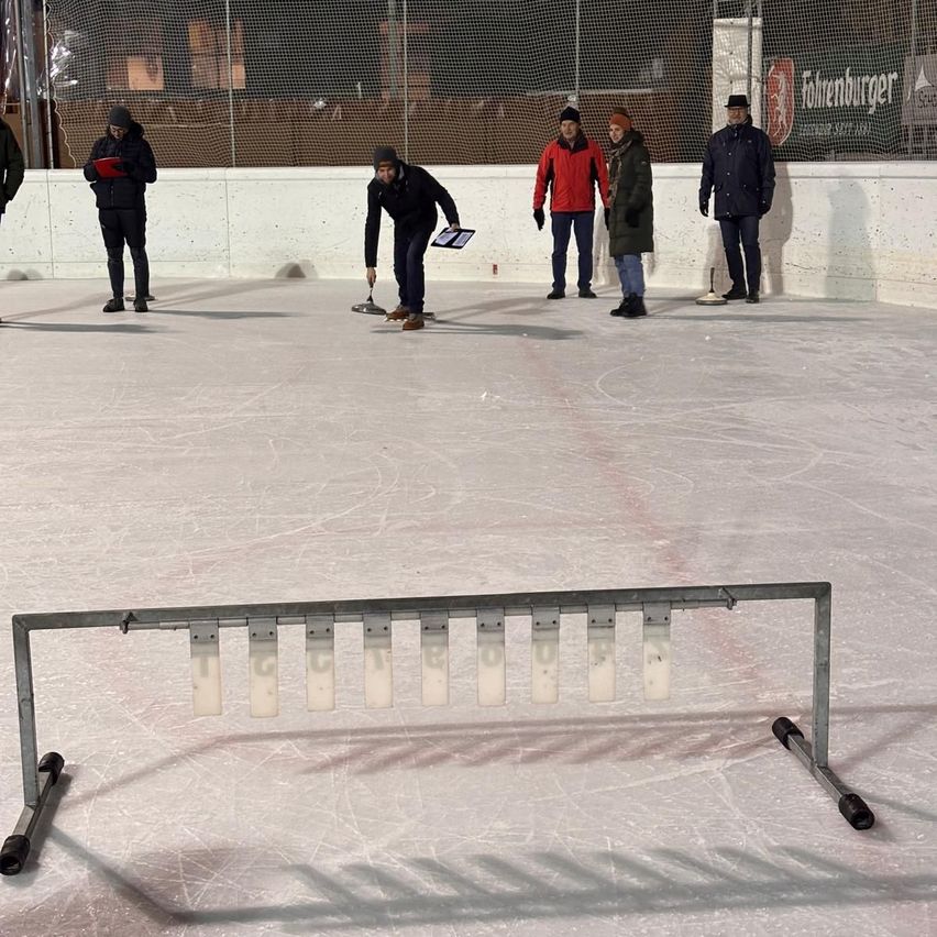 Eine Gruppe von Menschen ist auf einer Eisbahn. Eine Person beugt sich vor und hält einen Stock. Hinter ihm stehen drei weitere Personen. Eine Metallbarriere ist auf der Eisbahn platziert.