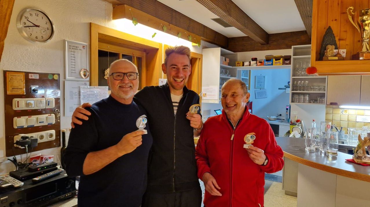 Drei Männer stehen lächelnd zusammen. Der Mann auf der linken Seite trägt eine Brille und hält einen Pokal. Der Mann in der Mitte hat seinen Arm um den linken Mann gelegt und hält einen Pokal. Der Mann auf der rechten Seite trägt eine rote Jacke und hält einen Pokal. Sie sind in einem Raum mit Holzbalken und Regalen.