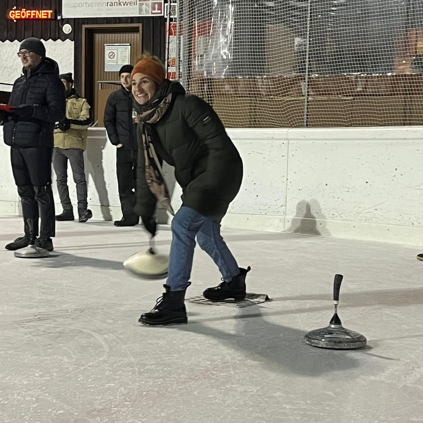 Eine Frau steht auf einer Eisbahn und wirft einen Curlingstein, während andere im Hintergrund zusehen. Die Bahn ist von einer weißen Barriere umgeben, auf der ein Schild mit der Aufschrift 'Rankweil' steht.