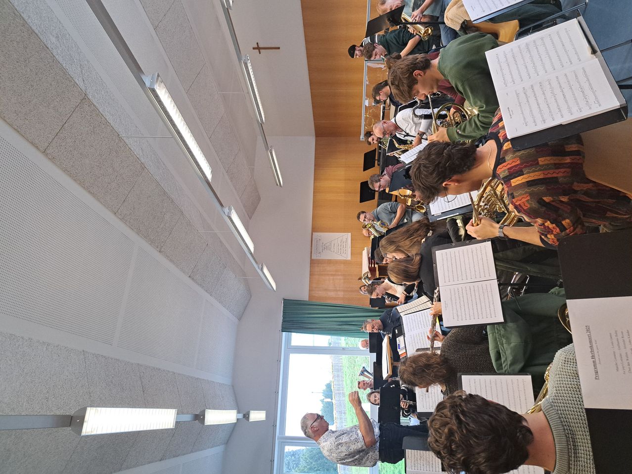 Ein Orchester probet in einem Raum mit Holzwänden, einem grünen Vorhang und weißen Deckenleuchten. Die Musiker konzentrieren sich auf ihre Instrumente, einige halten Notenblätter.