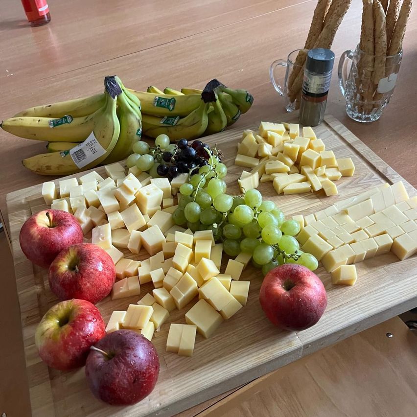 Ein Holzschneidebrett zeigt verschiedene Lebensmittel wie Käse, Trauben, Äpfel und Bananen. Brotsticks sind in zwei Glasbechern auf dem Tisch.