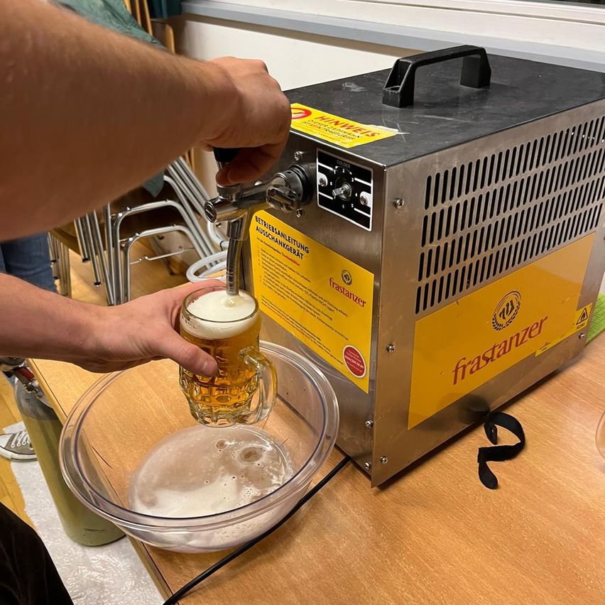 Eine Person füllt Bier aus einer Maschine in ein Glas auf einem Holztisch. Die Maschine hat ein gelbes Etikett mit dem Wort 'franstanzer'.
