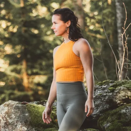 Eine Frau geht durch einen Wald und trägt ein orangefarbenes Tanktop und graue Leggings.