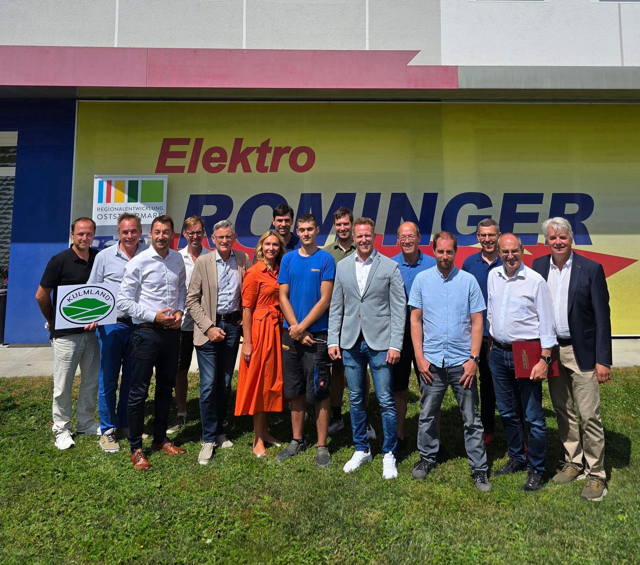 Eine Gruppe von Menschen steht vor einem Elektro Rominger Gebäude. Eine Person hält ein Schild mit dem Text 'KulmlAnd'. Sie sind alle formell gekleidet, lächeln und scheinen für ein Foto zu posieren.