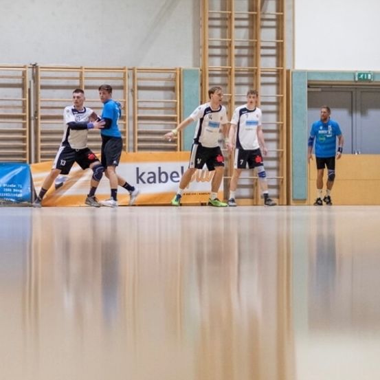 Fünf Männer spielen Futsal in einer Turnhalle. Vier stehen, einer fällt. Ein Werbebanner ist an der Wand hinter ihnen zu sehen.