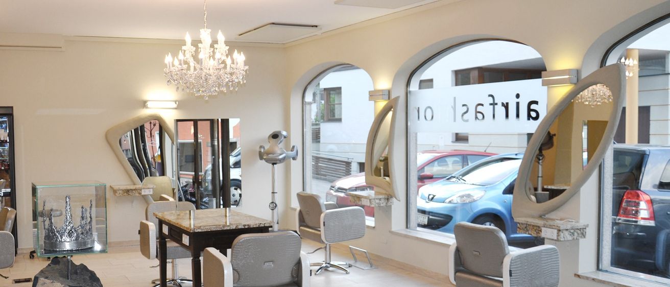 Ein Salon-Interieur mit mehreren Stühlen und einem Tisch. Eine Kristallleuchte und Spiegel sind an der Wand angebracht. Draußen vor dem Salon sind Autos geparkt.