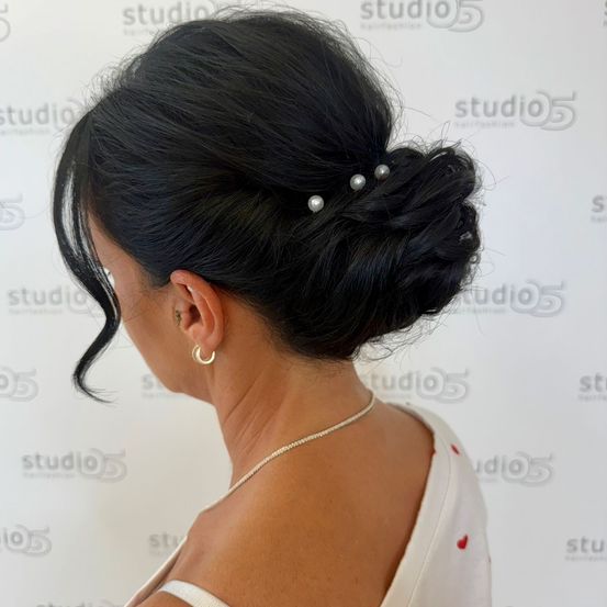 Eine Frau mit schwarzen Haaren in einem Dutt, geschmückt mit Perlennadeln, trägt ein weißes Kleid und eine Halskette. Sie steht vor einer weißen Wand mit Logos.