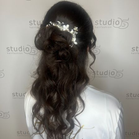 Eine Frau mit langen, gewellten Haaren in einem Dutt mit einem dekorativen Haarstück, von hinten in einem Studio gesehen.