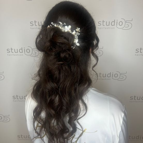 Eine Frau mit langen, gewellten Haaren in einem Dutt mit einem dekorativen Haarstück, von hinten in einem Studio gesehen.