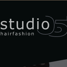 Ein schwarzes Bild zeigt das Logo von Studio 5 Hairfashion mit einem schlanken, modernen Design. Unter dem Logo ist ein verschwommenes Bild eines Salons mit einem Schreibtisch und einigen Objekten zu sehen.