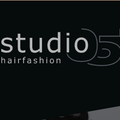 Ein schwarzes Bild zeigt das Logo von Studio 5 Hairfashion mit einem schlanken, modernen Design. Unter dem Logo ist ein verschwommenes Bild eines Salons mit einem Schreibtisch und einigen Objekten zu sehen.